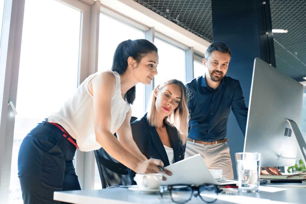 Three business people in a bright office are working together at a computer. A woman with dark hair in a white top is leaning over the desk, a blonde woman in a dark blazer is sitting in front of it, and a man with a beard in a blue shirt is standing next to it. They are all looking intently at the screen. There is a glass of water, a coffee cup and a pair of glasses on the desk.