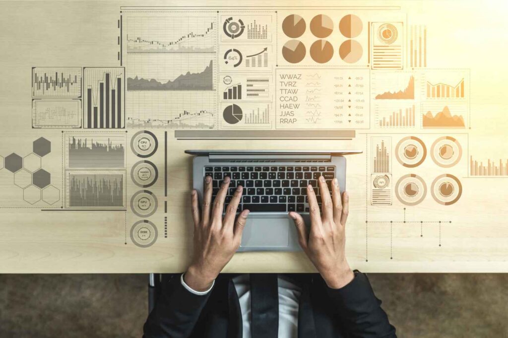 Aufsicht auf die Hände einer Person in Geschäftskleidung, die auf der Tastatur eines Laptops tippen. Der Laptop steht auf einem hellen Holztisch und ist ringsum von zahlreichen stilisierten, digital erscheinenden Diagrammen, Graphen und Daten-Visualisierungen umgeben, die auf oder über der Tischoberfläche schweben.