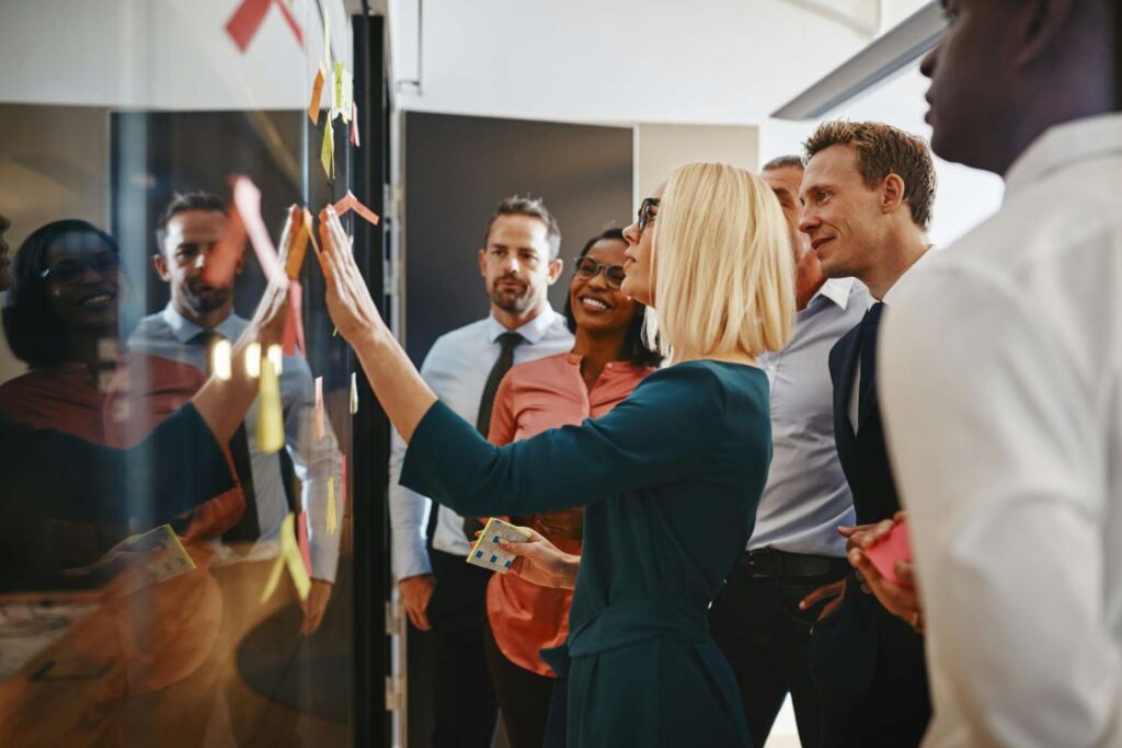 Eine Gruppe von mehreren Geschäftsleuten steht in einem modernen Büroraum um eine transparente Glaswand oder Tafel, die mit vielen bunten Haftnotizen beklebt ist. Eine Frau im Vordergrund, seitlich zur Kamera, befestigt gerade aktiv eine weitere Haftnotiz an der Wand, während ihre Kollegen ihr dabei zusehen, teilweise lächelnd und aufmerksam. Die Szene wirkt wie eine kollaborative Planungs- oder Brainstorming-Sitzung.