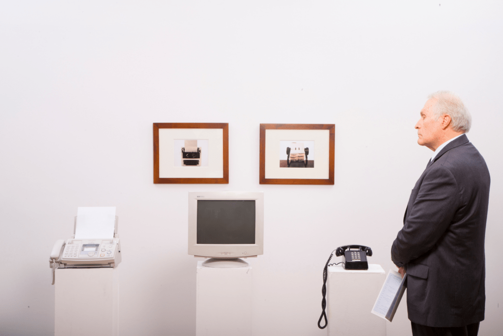 An older man in a suit stands in front of a white wall and looks at an exhibition of old office equipment: a fax machine, a CRT monitor and a rotary dial telephone. Framed pictures of office objects hang on the wall.