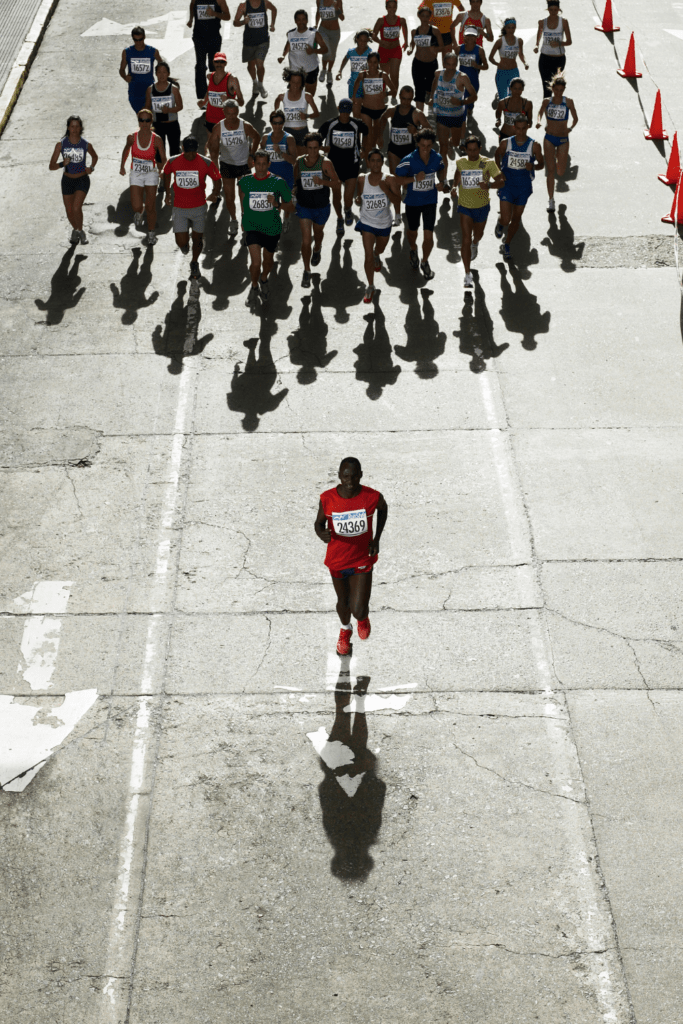Aerial view of a marathon runner in a red jersey running away from a group of runners. Long shadows of the runners fall on the asphalt of the road.