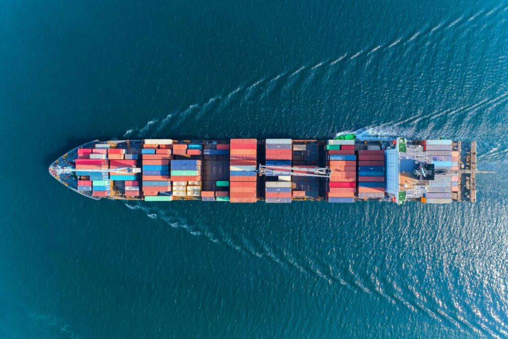 Luftbild eines großen Containerschiffs, das beladen mit bunten Containern über das blaue Meer fährt.